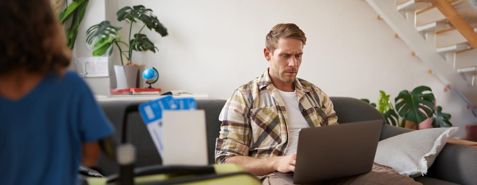 Homem pesquisa em um notebook sentado no sofá ao lado de uma mala aberta e de um globo terrestre.